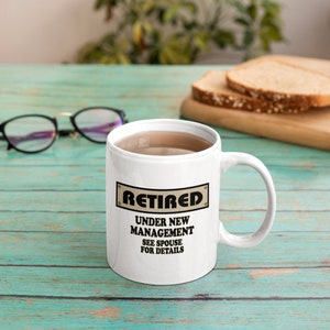 Op de afbeelding: Witte keramische koffiemok met een zwarte en bruine afbeelding met de tekst "Met pensioen Onder Nieuw Management Zie Echtgenoot Voor Details".