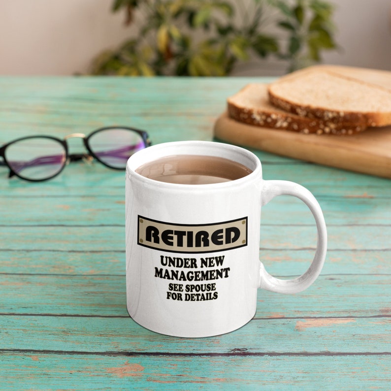 Op de afbeelding: Witte keramische koffiemok met een zwarte en bruine afbeelding met de tekst "Met pensioen Onder Nieuw Management Zie Echtgenoot Voor Details".