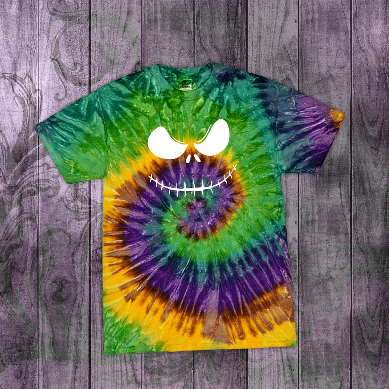 May include: A tie-dye t-shirt with a spiral pattern in green, yellow, purple, and brown. The shirt features a white graphic of a spooky face with large eyes and a stitched grin, perfect for Halloween.
