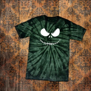 May include: A dark green tie-dye t-shirt with a white, cartoonish face design. The face features large, oval eyes and a stitched-like grin. The shirt is laid flat on a wooden surface with a floral pattern.