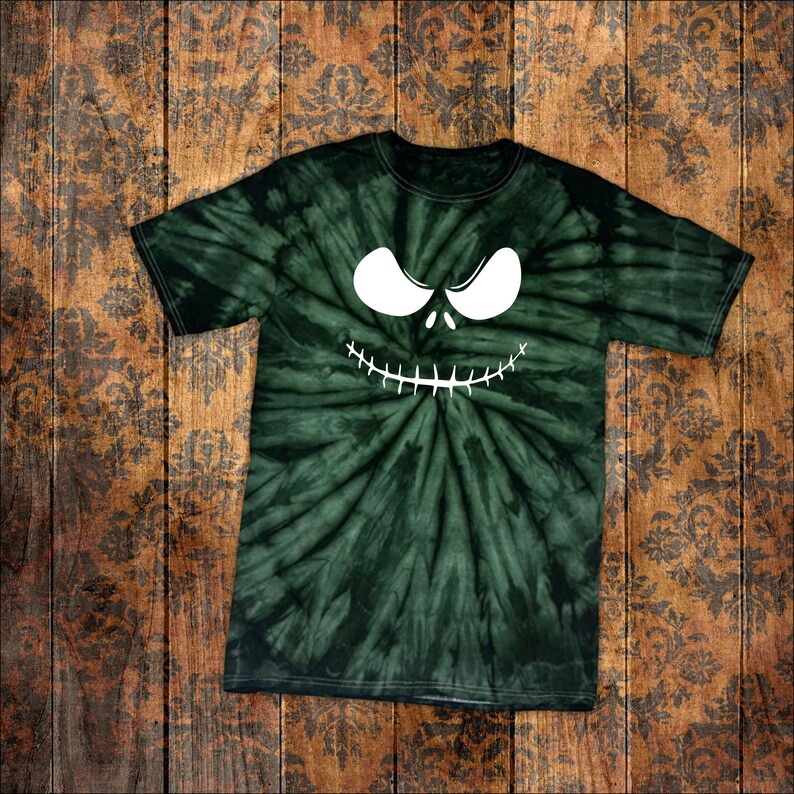 May include: A dark green tie-dye t-shirt with a white, cartoonish face design. The face features large, oval eyes and a stitched-like grin. The shirt is laid flat on a wooden surface with a floral pattern.