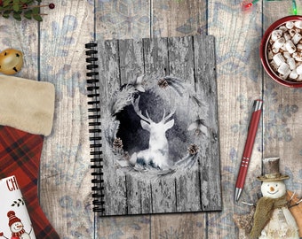 May include: A spiral-bound notebook featuring a gray wood-plank background with a central circular wreath design. The wreath surrounds a white deer silhouette and includes pinecones and evergreen branches. The cover has a rustic, weathered aesthetic.