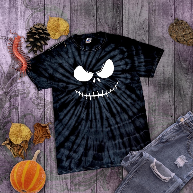 May include: Black tie-dye t-shirt featuring a white Jack Skellington face design. The shirt is paired with blue denim jeans. The image is set against a rustic wooden background with fall-themed decorations.