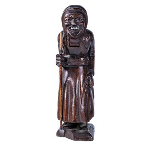 May include: A wooden nutcracker figurine of a woman with a long brown dress, holding a stick in her right hand. She has a carved face with a wide smile and a long, white beard.