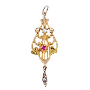 May include: A gold pendant with an ornate design. It features a central ruby-colored stone, an opal, and small pearls. The pendant has leaf accents and a delicate, elongated shape, perfect for a necklace.