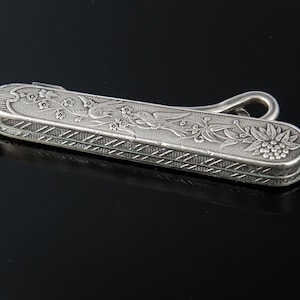 May include: A silver-colored, rectangular nail clipper with a decorative floral and bird design. The clipper has a curved handle and a textured, patterned edge. The design includes flowers, leaves, and a bird.