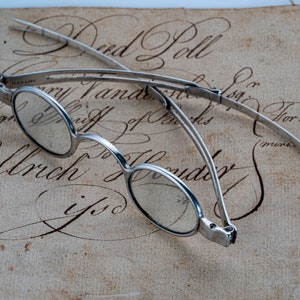 May include: A pair of antique silver-toned folding eyeglasses with round lenses on a piece of old paper with handwritten text that reads "Bond Loll" and "Mary Fandarar for 42.A."