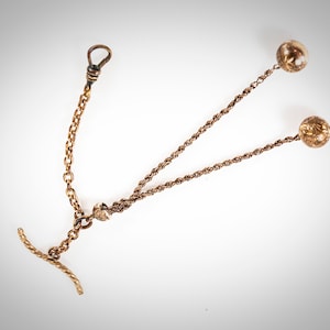 May include: A gold pocket watch chain with a T-bar and two decorative balls at the ends.