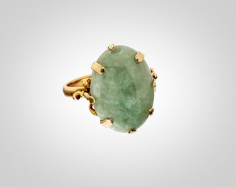 May include: A gold ring with a large, oval-shaped green jade stone set in a prong setting. The ring has a delicate, ornate design.