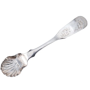May include: A silver sugar spoon with a shell-shaped bowl and a handle with engraved floral and initial designs. The spoon is made of a shiny, reflective metal and is set against a white background.
