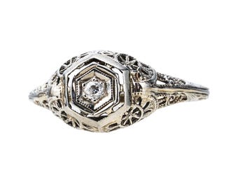 May include: A vintage silver ring with a central hexagonal setting. The setting holds a small, round diamond. The ring features intricate filigree detailing and floral patterns on the band and shoulders, creating an antique aesthetic.