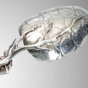 May include: A silver serving spoon with a detailed floral design on the bowl and a branch-like handle. The spoon's bowl has a textured, layered appearance, and the handle features intricate branch and twig details.
