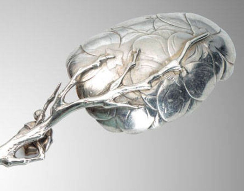 May include: A silver serving spoon with a detailed floral design on the bowl and a branch-like handle. The spoon's bowl has a textured, layered appearance, and the handle features intricate branch and twig details.