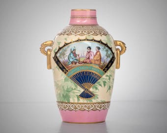 May include: A pink and white porcelain vase with gold accents and a painted scene of three figures in traditional clothing. The vase has two gold handles and a fan design.