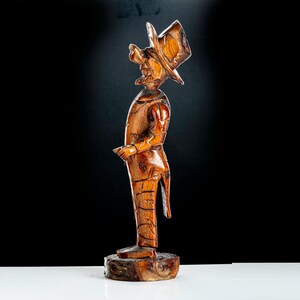May include: A wooden figurine of a man wearing a top hat and a suit. The figurine is carved in a rustic style and has a brown finish.