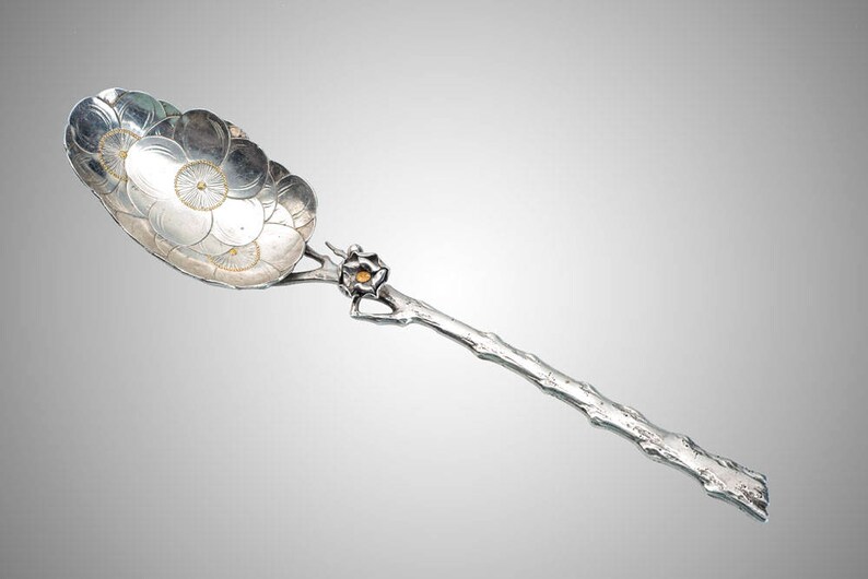 May include: A silver serving spoon with a floral design on the bowl. The handle is shaped like a branch, and the spoon has a small, gold-colored accent. The spoon is likely used for serving food or as a decorative piece.