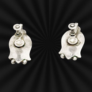 May include: A pair of silver tone clip-on earrings shaped like a flower with a screw back closure. The earrings are marked "SILVER" on the back.