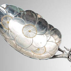 May include: A decorative silver serving spoon with a floral design. The spoon's bowl is composed of overlapping silver petals, with gold accents in the center. The handle features a stylized floral motif.