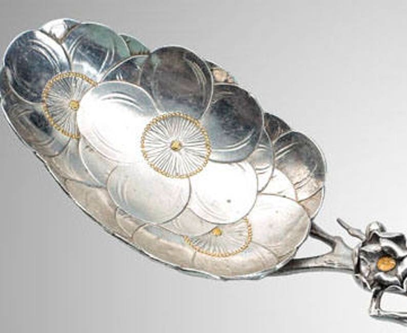 May include: A decorative silver serving spoon with a floral design. The spoon's bowl is composed of overlapping silver petals, with gold accents in the center. The handle features a stylized floral motif.