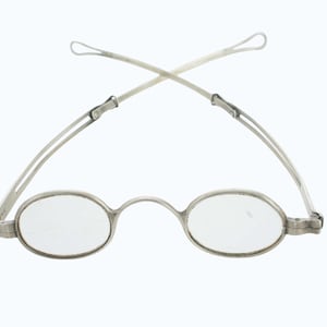 May include: A pair of silver-toned metal spectacles featuring oval-shaped lenses and a thin bridge. The frame includes hinged, slender temples that extend upward and cross each other, terminating in small, rounded loops at the ends.