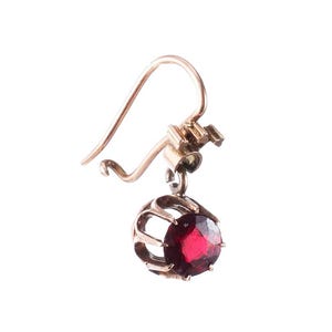 May include: A single gold-colored earring with a deep red gemstone. The gemstone is round and set within a cage-like design. The earring has a hook closure.
