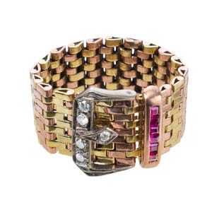 May include: A wide, multi-tone ring with a buckle design. The ring features gold, rose gold, and copper-colored metal links. The buckle is silver-toned with clear gemstones and a row of red gemstones.