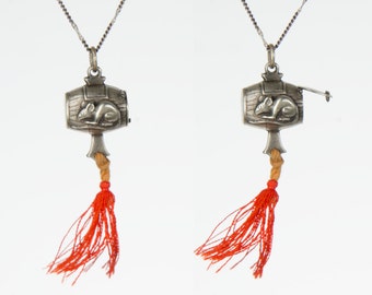 May include: A silver pendant necklace with a small, detailed mouse figurine inside a barrel-shaped compartment. The pendant has a red tassel attached to it.