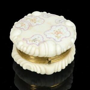 May include: A round, off-white trinket box with a gold-colored metal band. The lid features floral designs in pink, gold, and white, with a scalloped edge. The box has a textured, ruffled base and lid.