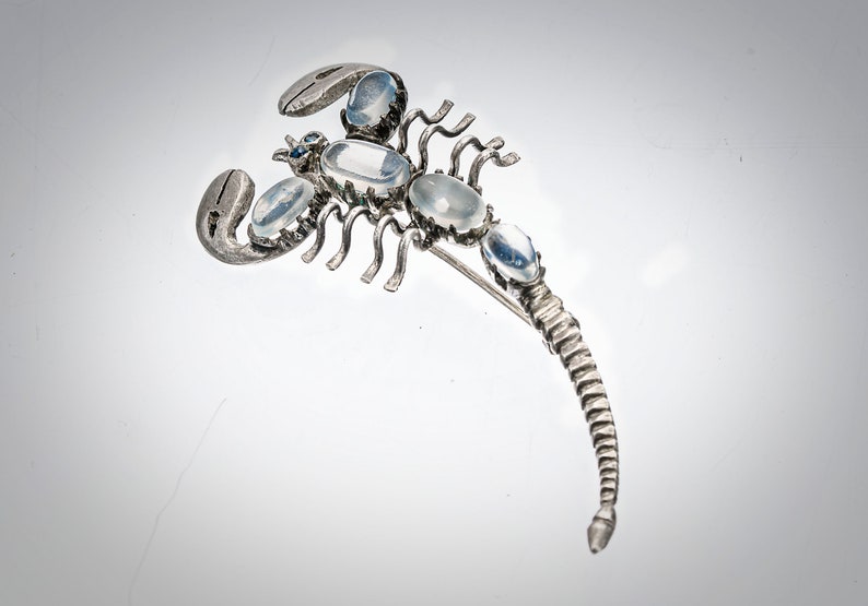 May include: A silver scorpion brooch with clear moonstone accents. The scorpion's tail is curled up and the brooch has a pin on the back.