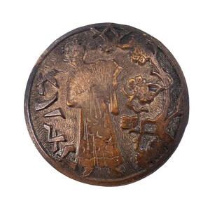 May include: A round, bronze-colored decorative item with an embossed design. The design features a figure holding a fan, with Japanese characters and floral elements. The item has a detailed, textured surface and is set against a white background.