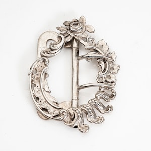May include: Ornate silver-colored buckle with floral and scrollwork details. The buckle features a central bar and a decorative rose at the top. The design is symmetrical and detailed, with a vintage aesthetic.