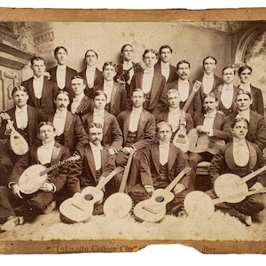 May include: A black and white photograph of a group of men in formal attire holding musical instruments. The men are standing and sitting in a room with a fireplace in the background. The photo is labeled "Lafayette College Class of 1897".