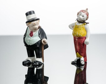 May include: Two vintage ceramic figurines. One figure is a man in a black suit, top hat, and holding a cane, with a cigar in his mouth. The other is a woman in a yellow top and red skirt, with a head covering.
