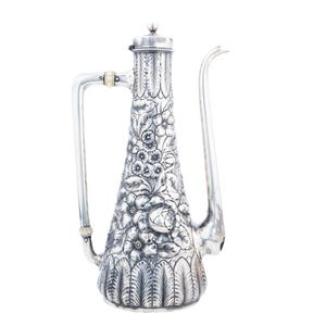 May include: A tall silver coffee pot featuring an intricate embossed floral pattern across the body, a long slender spout, a hinged lid with a small finial, and a large handle with light-colored accents at the top and bottom connection points.