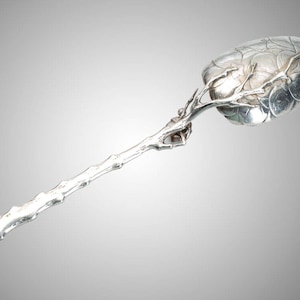 May include: A silver serving spoon with a unique design. The spoon's bowl has a leaf-like pattern, and the handle is shaped like a tree branch. The spoon is made of a shiny, reflective metal.