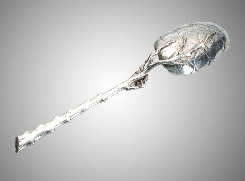 May include: A silver serving spoon with a unique design. The spoon's bowl has a leaf-like pattern, and the handle is shaped like a tree branch. The spoon is made of a shiny, reflective metal.