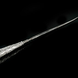 May include: A silver shoe horn with an ornate, engraved handle. The handle is decorated with a swirling floral pattern.