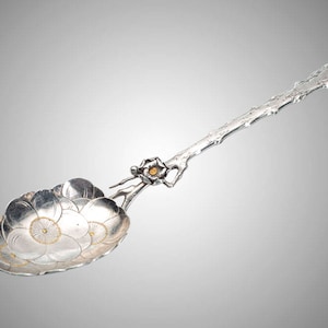 May include: A silver spoon with a floral design. The spoon's bowl is shaped like a flower with delicate petals and gold accents. The handle is designed to look like a branch, with a small flower detail near the bowl.