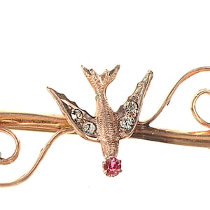 May include: A gold-toned brooch featuring a stylized bird with a red gemstone on its chest, set on a delicate branch with leaves.