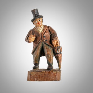 May include: A wooden figurine of a man wearing a top hat and a brown suit, holding an umbrella. The figurine is standing on a wooden base.