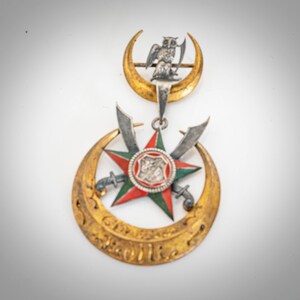 May include: A gold and silver pendant with a red, green, and white star, two swords, and a crescent moon. The pendant features an inscription in Arabic script and an owl perched on a crescent moon.