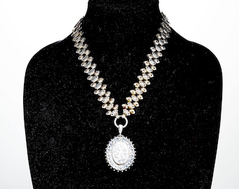 Pode incluir: Um colar de corrente de prata com um pingente de medalhão de prata. A corrente é feita de elos entrelaçados e tem um acabamento dourado. O medalhão é oval e tem um design detalhado.