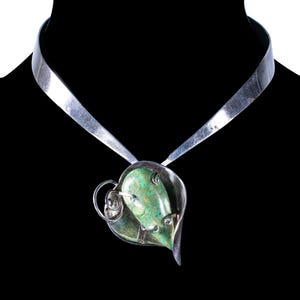 May include: A silver-toned necklace with a leaf-shaped pendant. The pendant features a green stone and decorative silver wire accents. The necklace has a wide, flat collar design, and the background is black.