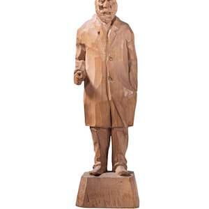 May include: A wooden carving of a person wearing a hat and coat, standing on a wooden base. The carving is signed "A. Wipfler".