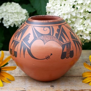 May include: A terracotta-colored ceramic vase with a rounded body and a wide opening. The vase is adorned with a black geometric design featuring stylized shapes and a heart-like motif. Yellow flowers and white blooms are in the background.