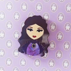 Puede incluir: Broche de mujer de pelo oscuro con vestido morado y azul. El broche tiene un círculo dorado en el vestido. El fondo es de color lila claro con un patrón blanco repetido.