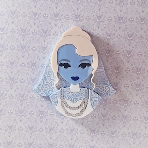 Op de afbeelding: Een blauwhuidige bruidachtige figuur met een witte sluier en jurk, versierd met een parelketting. De figuur heeft zwarte wimpers en donkerblauwe lippen. De achtergrond heeft een herhalend patroon van sierlijke ontwerpen.