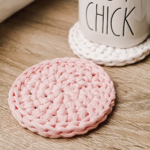 May include: Two crocheted coasters, one white and one pink. The pink coaster is in focus and the white coaster is out of focus in the background. The white coaster is under a white ceramic mug with the text "HOT CHICK" printed on it.
