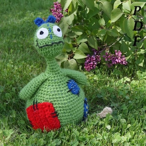 May include: A green crocheted monster toy with blue eyes, a red patch, and a blue patch. The toy is sitting on a bed of green grass.