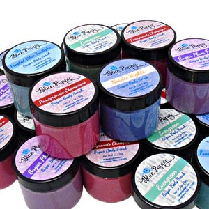 May include: A collection of small, round jars of sugar body scrub with black lids and colorful labels. The labels feature the brand name "Blue Poppy" and various scent names like "Pomegranate Champagne" and "Evergreen".